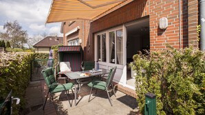 An outdoor patio with a table and chairs, a striped awning, and a brick house. - Four-star accommodation in Friesenhörn, in a quiet, south-facing location, right on the dike (Horumersiel)
