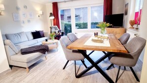 A modern living room with a wooden dining table, grey sofas, and a large window with red curtains. - Four-star accommodation in Friesenhörn, in a quiet, south-facing location, right on the dike (Horumersiel)