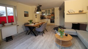 A modern living room with a dining area, a kitchen in the background, and a grey sofa with a small table in the foreground. - Four-star accommodation in Friesenhörn, in a quiet, south-facing location, right on the dike (Horumersiel)