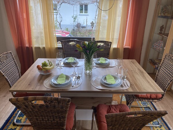 Une table à manger dressée pour quatre personnes, avec des chaises en osier, un vase de fleurs et un saladier rempli de fruits.