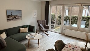 Uma sala de estar com um sofá verde, uma poltrona cinza, uma mesa de centro redonda e uma mesa de jantar com toalha de mesa florida.