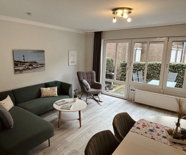 A living room with a green sofa, a grey armchair, a round coffee table, and a dining table with a floral tablecloth. - Four star oasis in Friesenhörn, quiet location, right on the dike (Horumersiel)