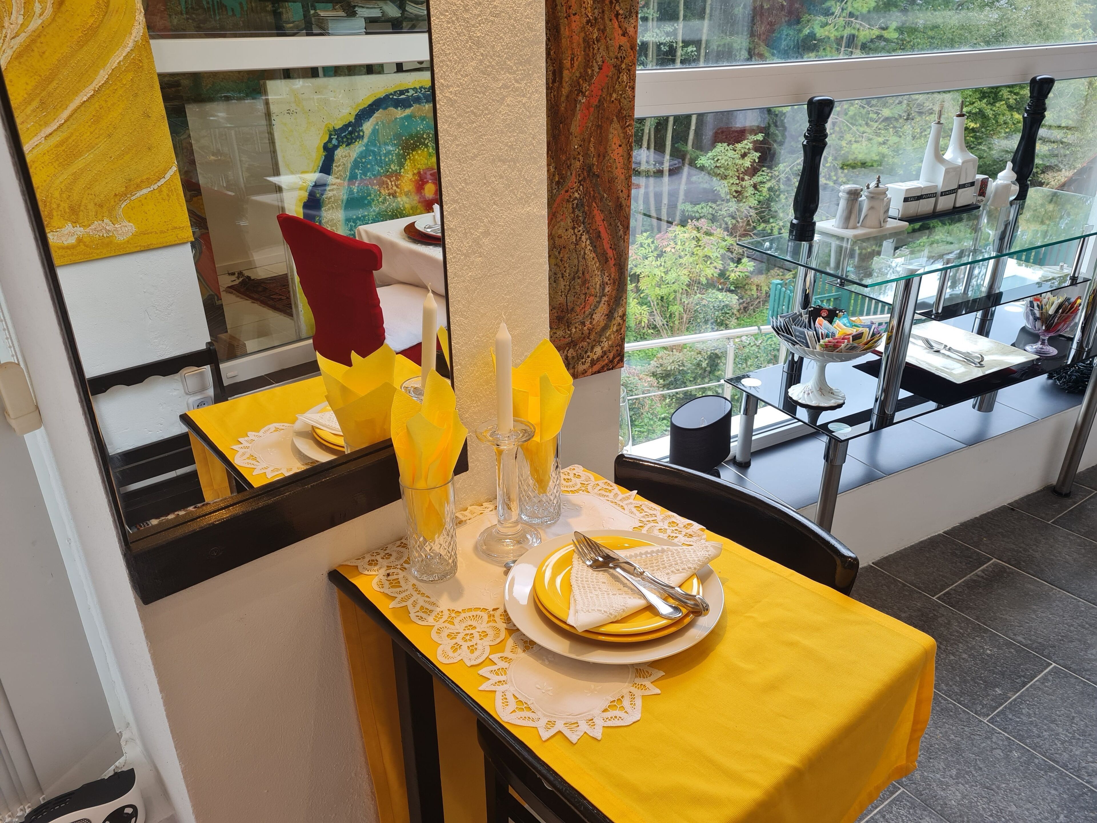 Un comedor con mantel amarillo, copitos de encaje blanco y ventana de vidrio con vista a un jardín.