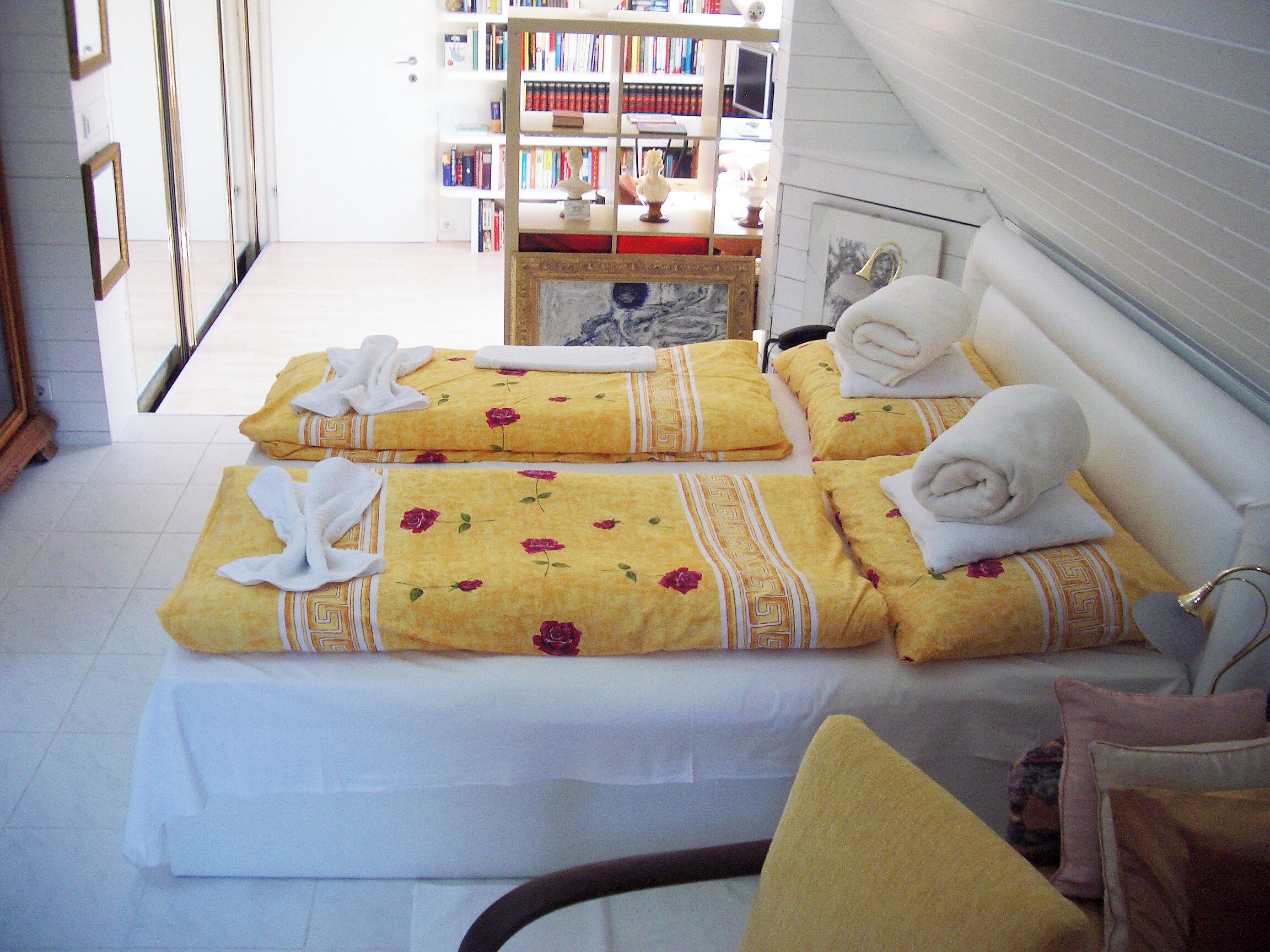 A hotel room with a bed, a chair, and a bookshelf in the background.