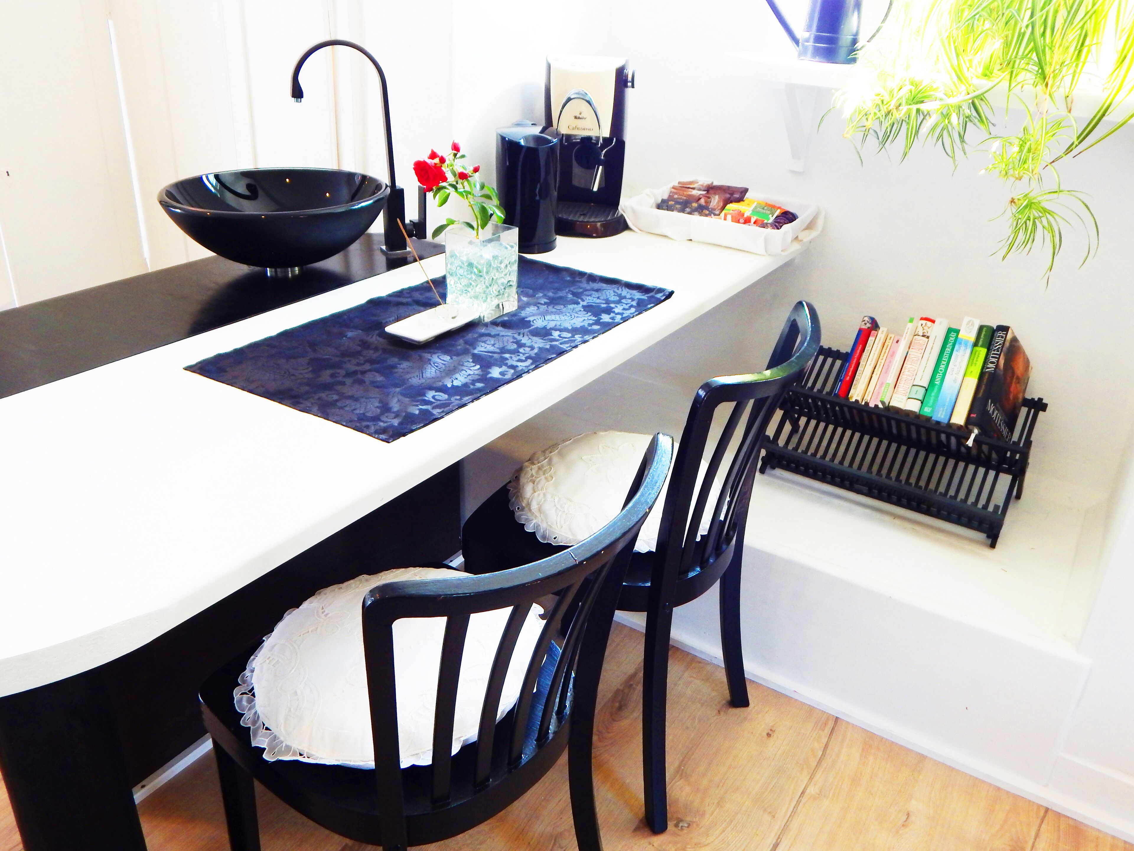 Uma sala de jantar com uma mesa branca, cadeiras pretas, uma tigela preta, uma máquina de café e uma estante de livros.