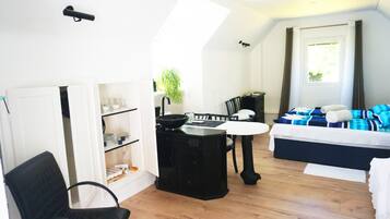 Kompakti hotellihuone, jossa on pieni keittiö, ruokailutila, sinisillä tekstiileillä varustettu sänky ja suuri ikkuna, jossa on verhot.