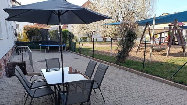 Une terrasse extérieure avec une table et des chaises, un barbecue et, au fond, un ensemble de jeux pour enfants avec une balançoire.