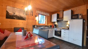 A wooden interior with a dining table set for two, a kitchen with modern appliances, and a window with a view of a mountain.