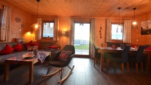 A cozy wooden cabin interior with a dining area, a rocking chair, and a sofa with red cushions.