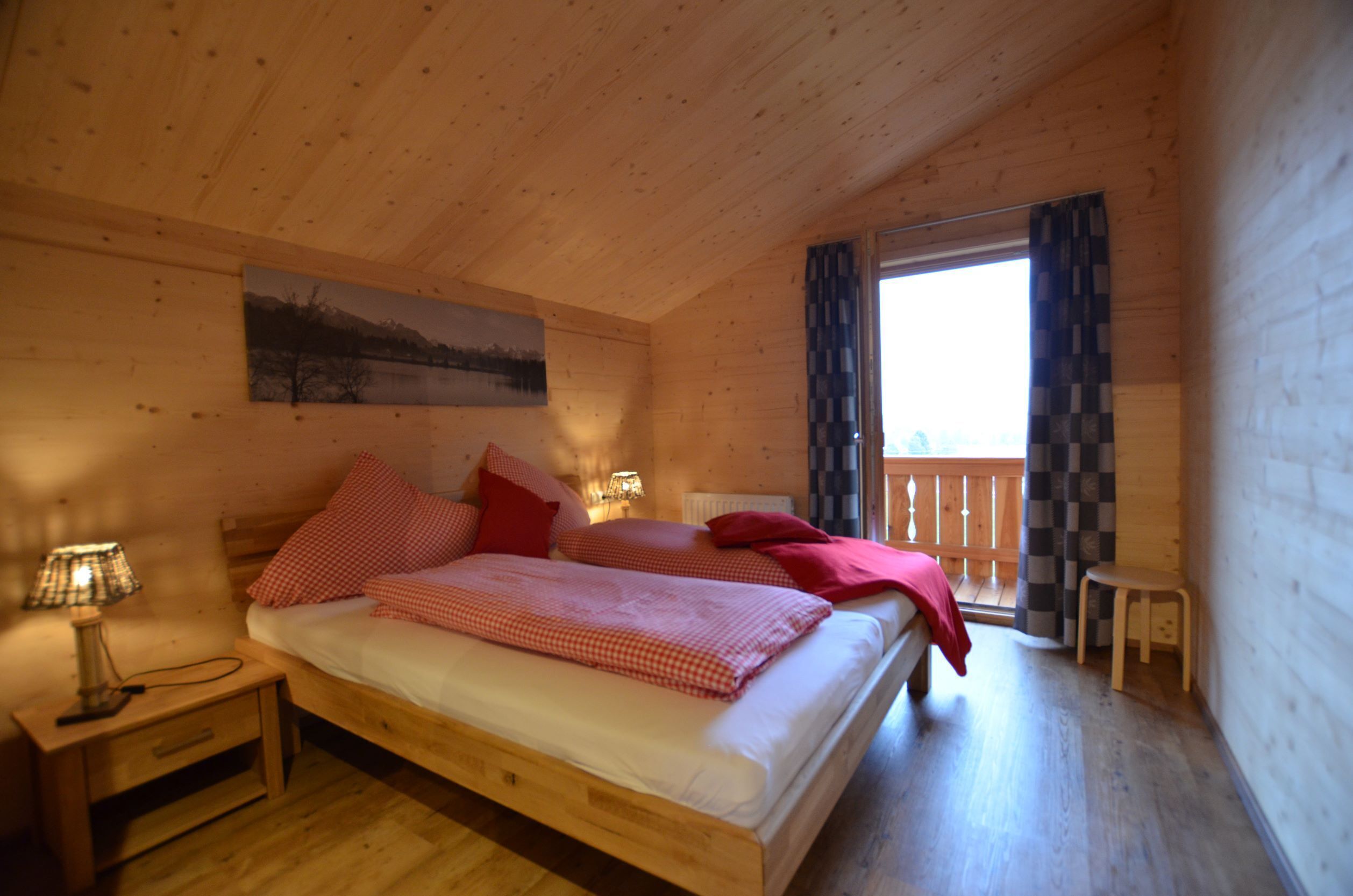 A wooden cabin bedroom with a bed, nightstands, a small table, and a view of the outdoors.