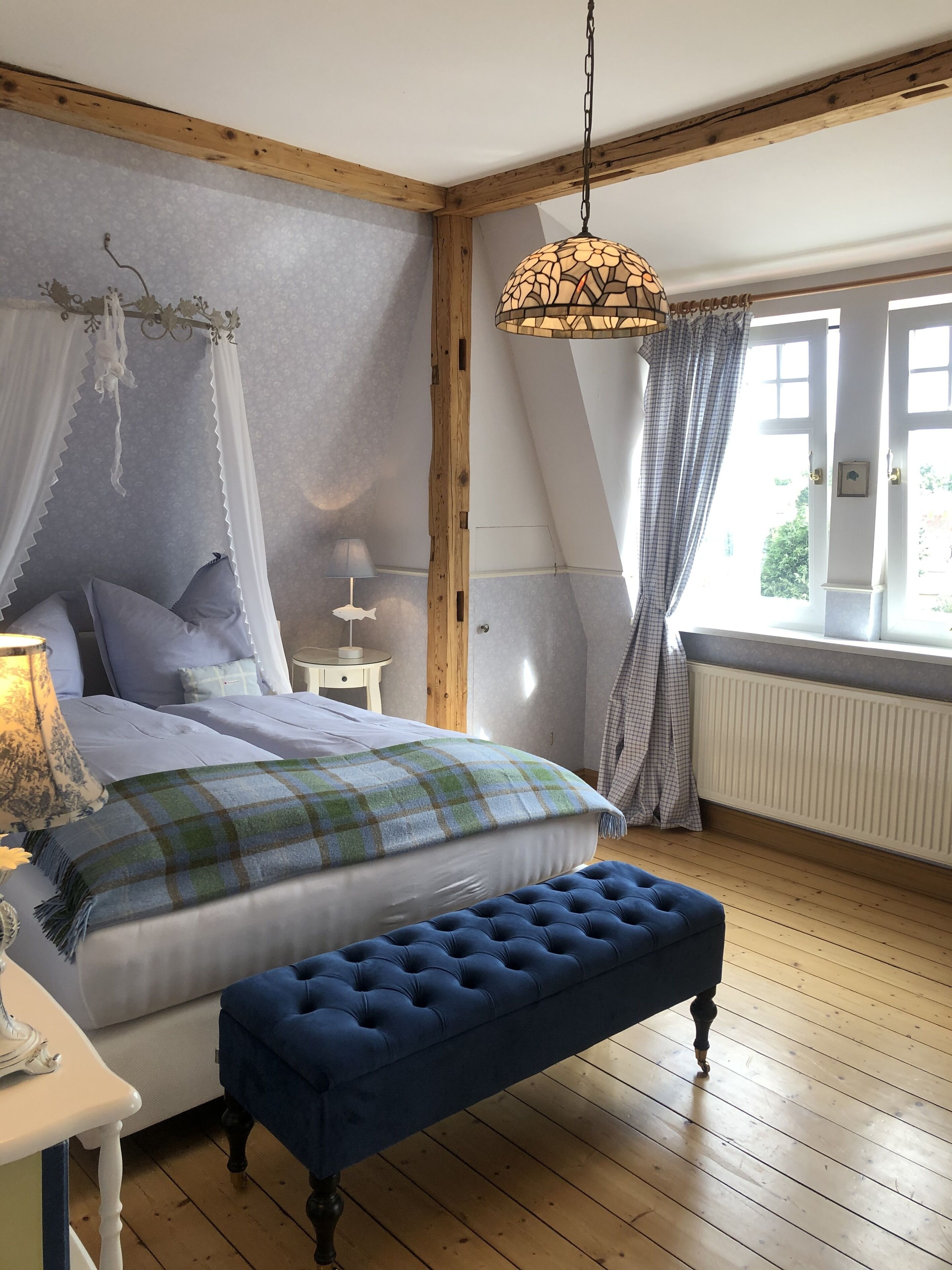 Une chambre à coucher comprenant un lit, un banc, une table de chevet, une lampe, une fenêtre avec des rideaux et un luminaire suspendu.