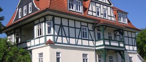 A large, two-story building with a symmetrical facade, featuring a mix of white and dark timber framing, multiple windows, and a red-tiled roof.