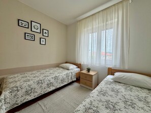 Un dormitorio con dos camas individuales, una mesita de noche, una pequeña mesa con una planta y una ventana con cortinas traslúcidas.