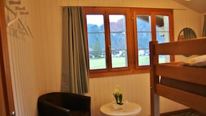 Una habitación con una cama, una silla, una mesita con un jarrón de flores y una ventana que ofrece vistas a una montaña y una casa.