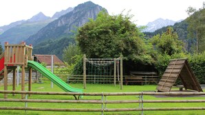 Un parque infantil con un tobogán y una red de escalada, rodeado por una valla de madera, con montañas al fondo.