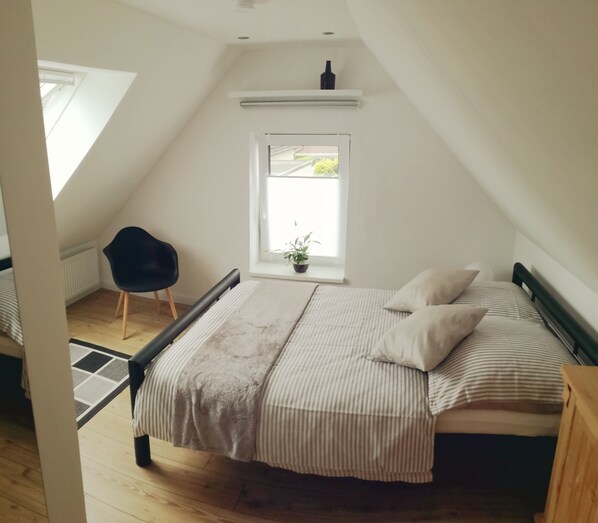 A bedroom with a sloped ceiling, a window with a view, a bed with striped bedding, and a chair.