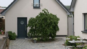 A building with a dark door, a window with a number, and a paved courtyard with a bench and potted plants.
