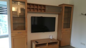 Un salon moderne avec un téléviseur à écran plat fixé au mur, des meubles en bois dotés de portes en verre et un petit meuble pour équipements multimédias placé en dessous.