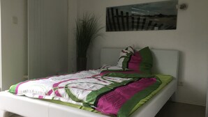 Un lit avec un cadre blanc, une couverture verte et rose, et un seul oreiller vert. Il y a une plante en pot à gauche du lit et un tableau accroché au mur au-dessus.