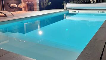 Ein Schwimmbad mit Holzterrasse, Kakteengestaltung und einem Tisch mit Stühlen.