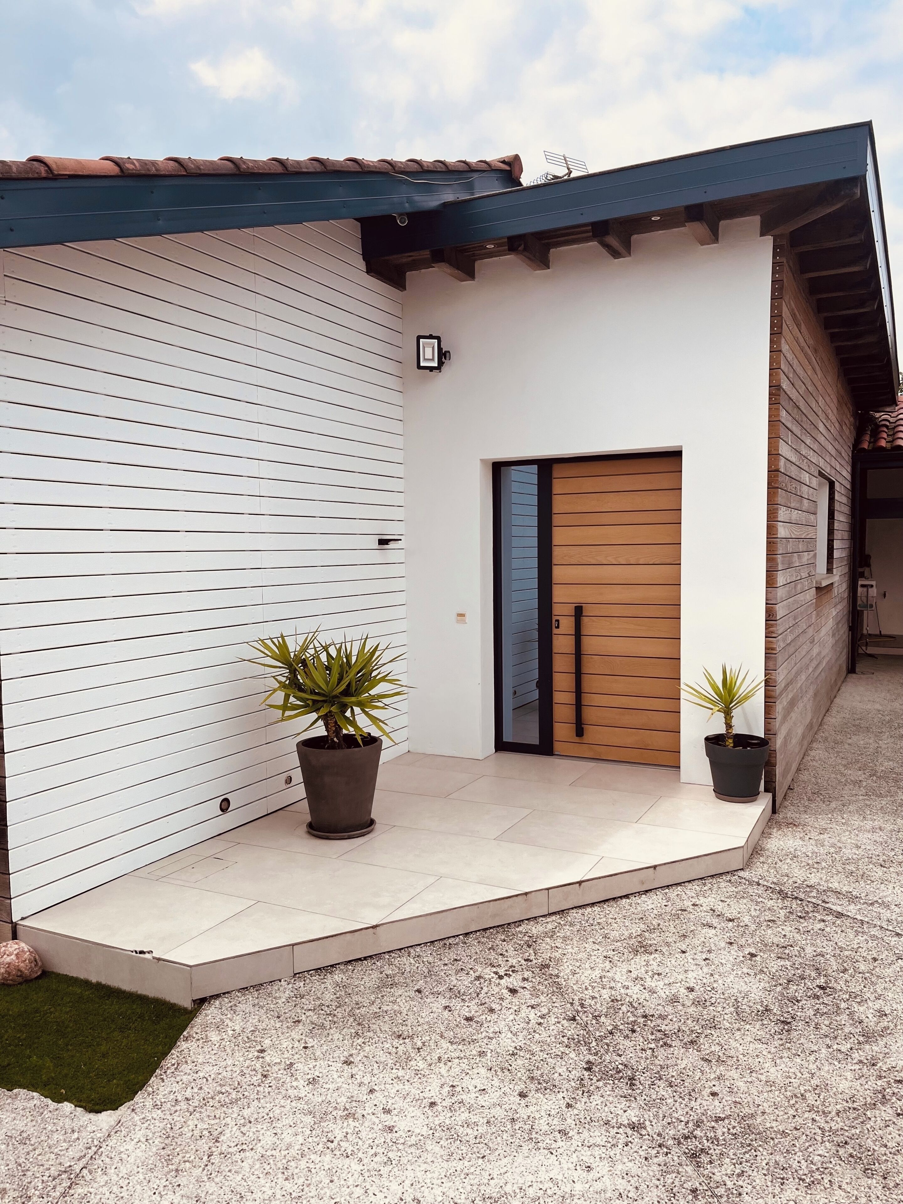 Een modern gebouw met een witte gevel, een houten deur en twee planten in potten op de veranda.