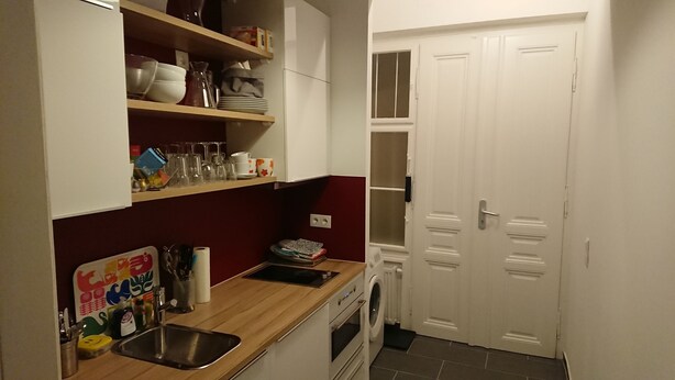 Una cocina compacta con un fregadero, estufa y horno. Hay repisas sobre la encimera con platos y vasos. Se ve una puerta blanca a la derecha.