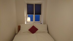 Una cama con ropa de cama blanca y un cojín rojo con lunares, una mesita de noche con lámpara y una ventana con persianas cerradas.