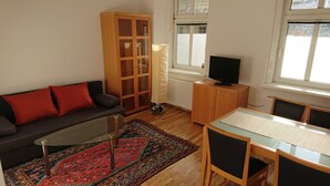 Una sala de estar con sofá, una mesa de centro de vidrio, un tapete estampado, un aparador de madera, un televisor y una ventana.