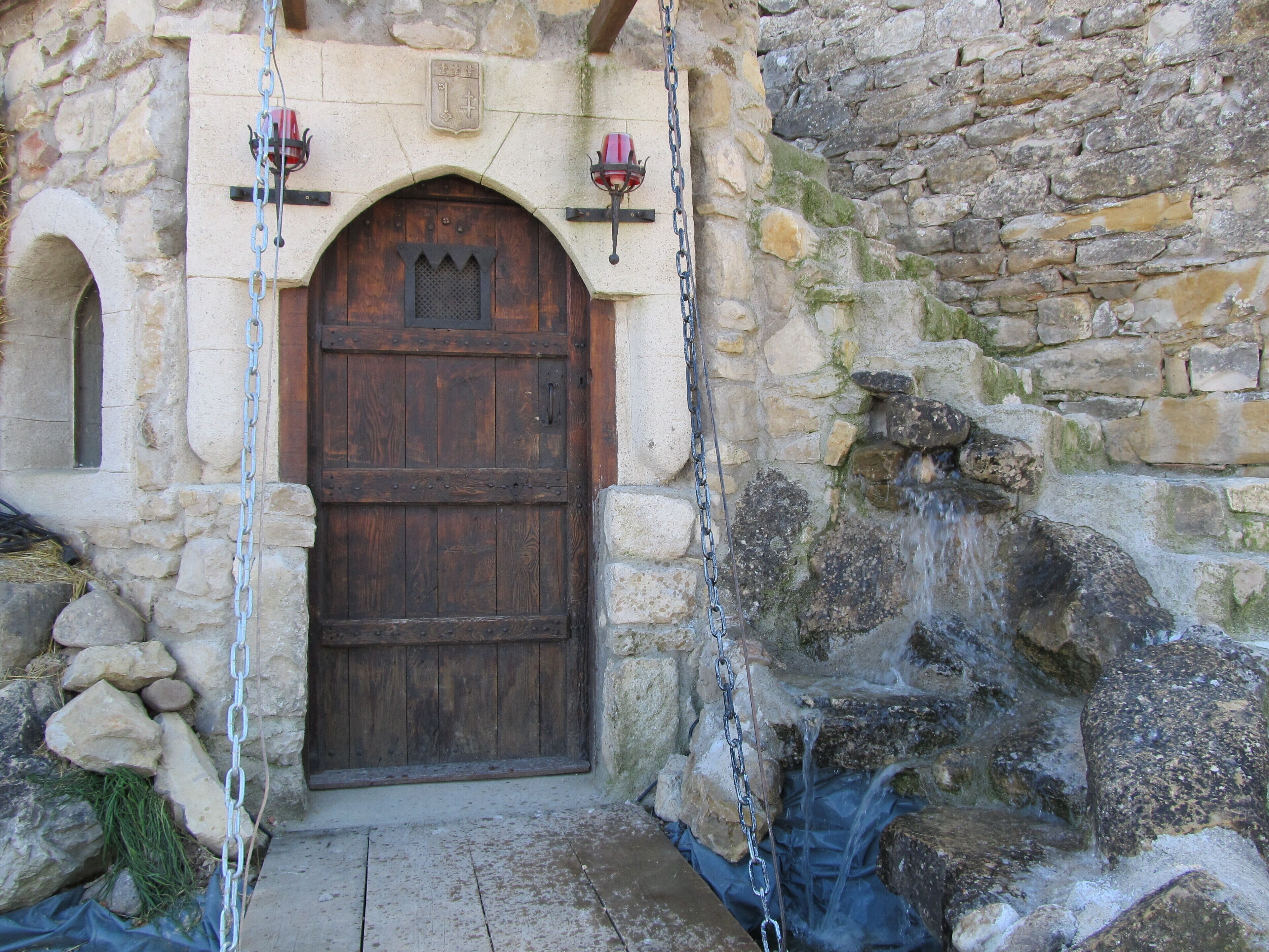 Un bâtiment en pierre avec une porte en bois, un mur en pierre agrémenté d’un élément décoratif aquatique et une chaîne suspendue à la porte.