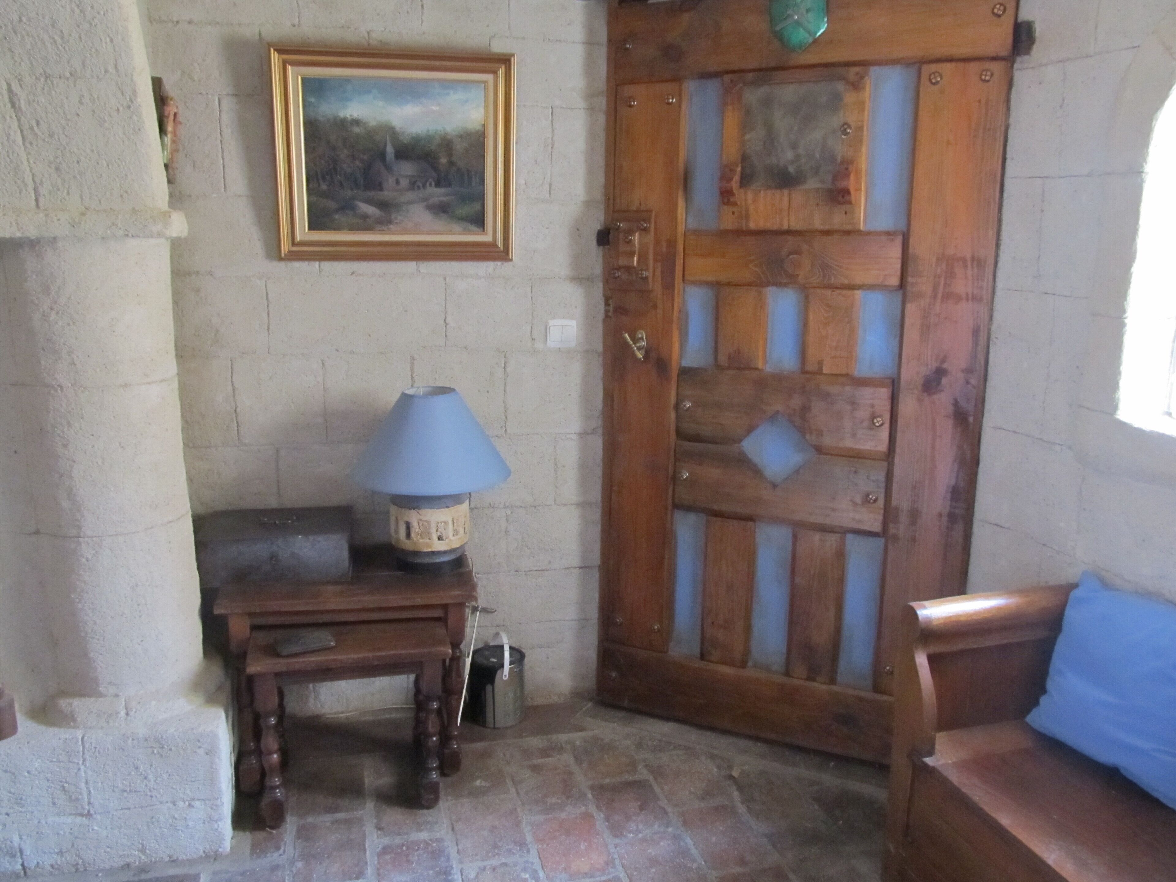 A room with a wooden door, a framed painting on the wall, a table with a lamp, and a bench.