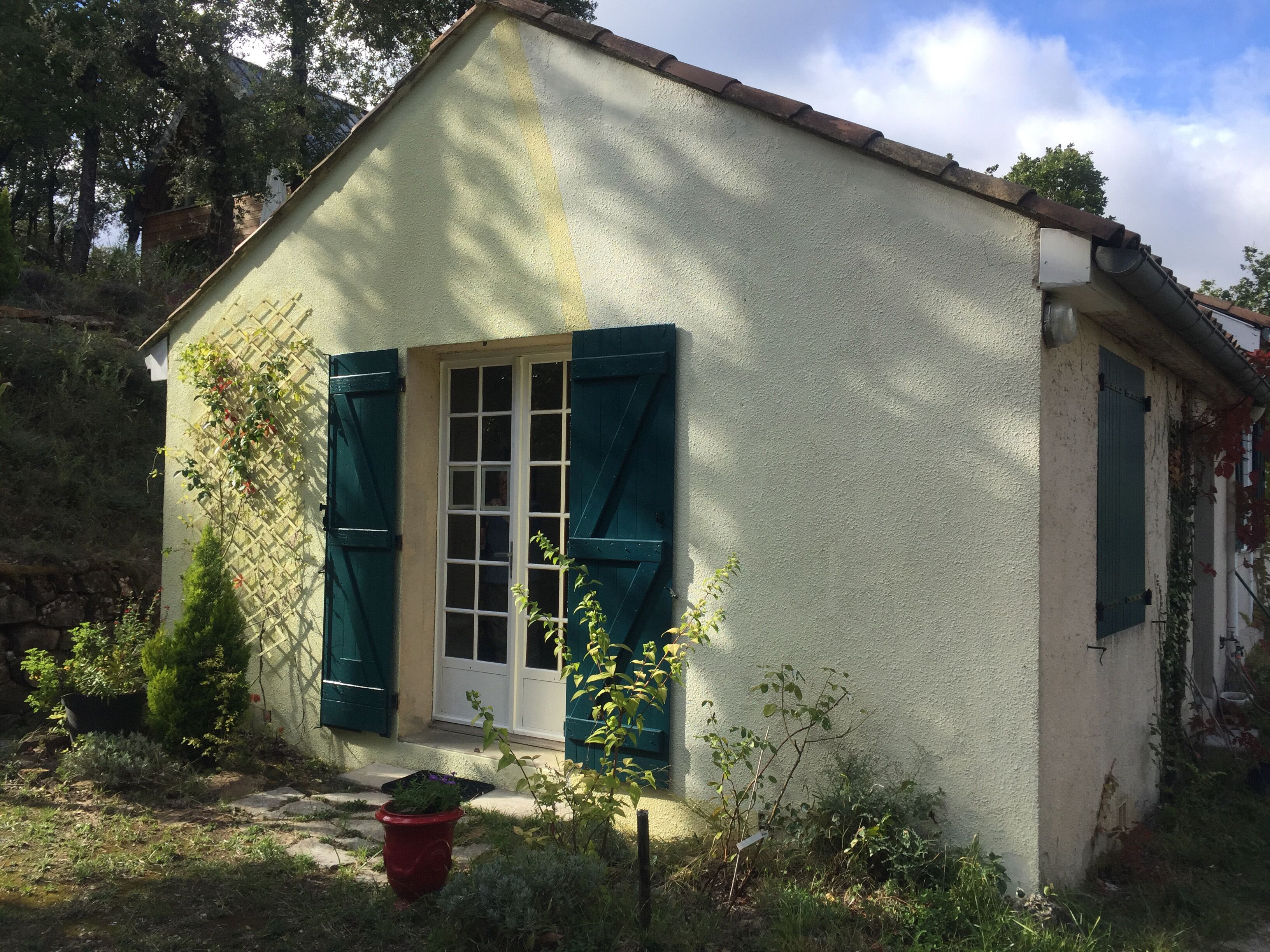 A small, single-story building with a white exterior and green shutters, surrounded by vegetation and a stone wall.