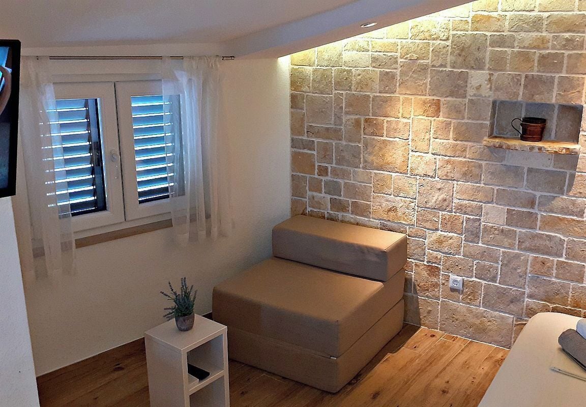 Ein gemütlicher Raum mit Steinwand, gepolstertem Sitzbrett, einem kleinen Regal mit einem Topf und einem Fenster mit Jalousien.