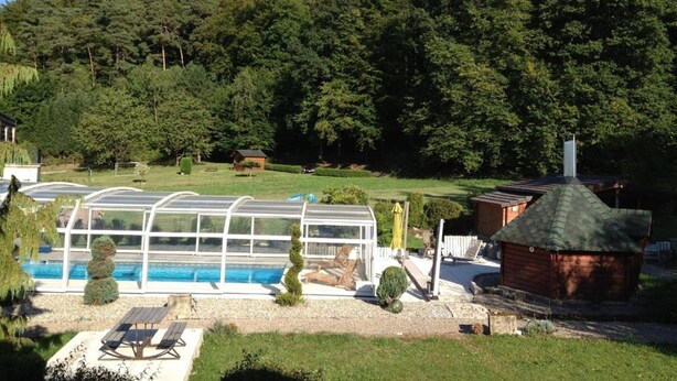 Un espace piscine doté d’une structure fermée par des parois de verre, d’une terrasse en bois et d’un petit bâtiment surmonté d’un toit végétalisé.