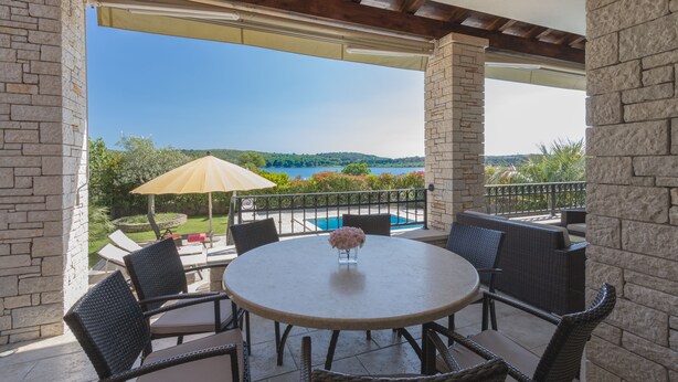A covered patio with a round table and chairs, overlooking a swimming pool and a scenic view of hills and water. - Luxury villa with heated pool by the sea in a quiet area (Pula - Pomer)