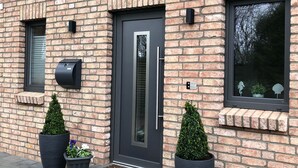 A modern brick building with a dark grey door, potted plants, and a window with decorative elements.