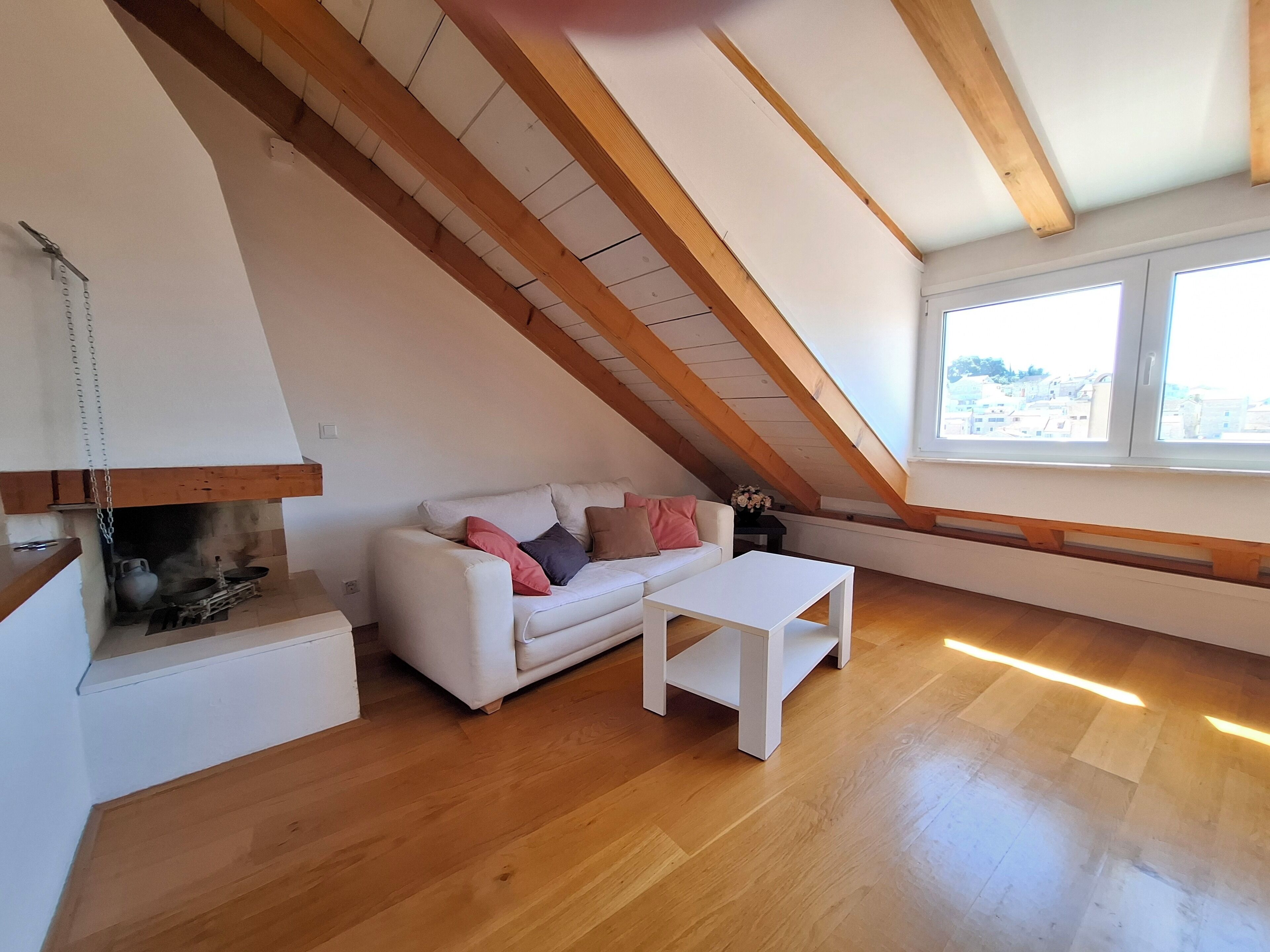 A loft-style room with a wooden ceiling, a white sofa with pillows, a small white table, and a view through a window.