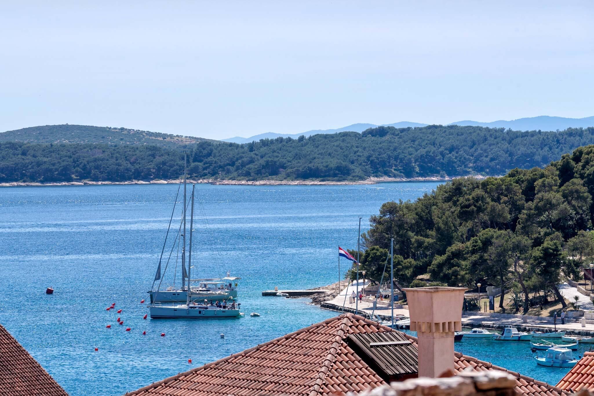 Vista costera con un velero, un muelle con barcos y montañas al fondo.