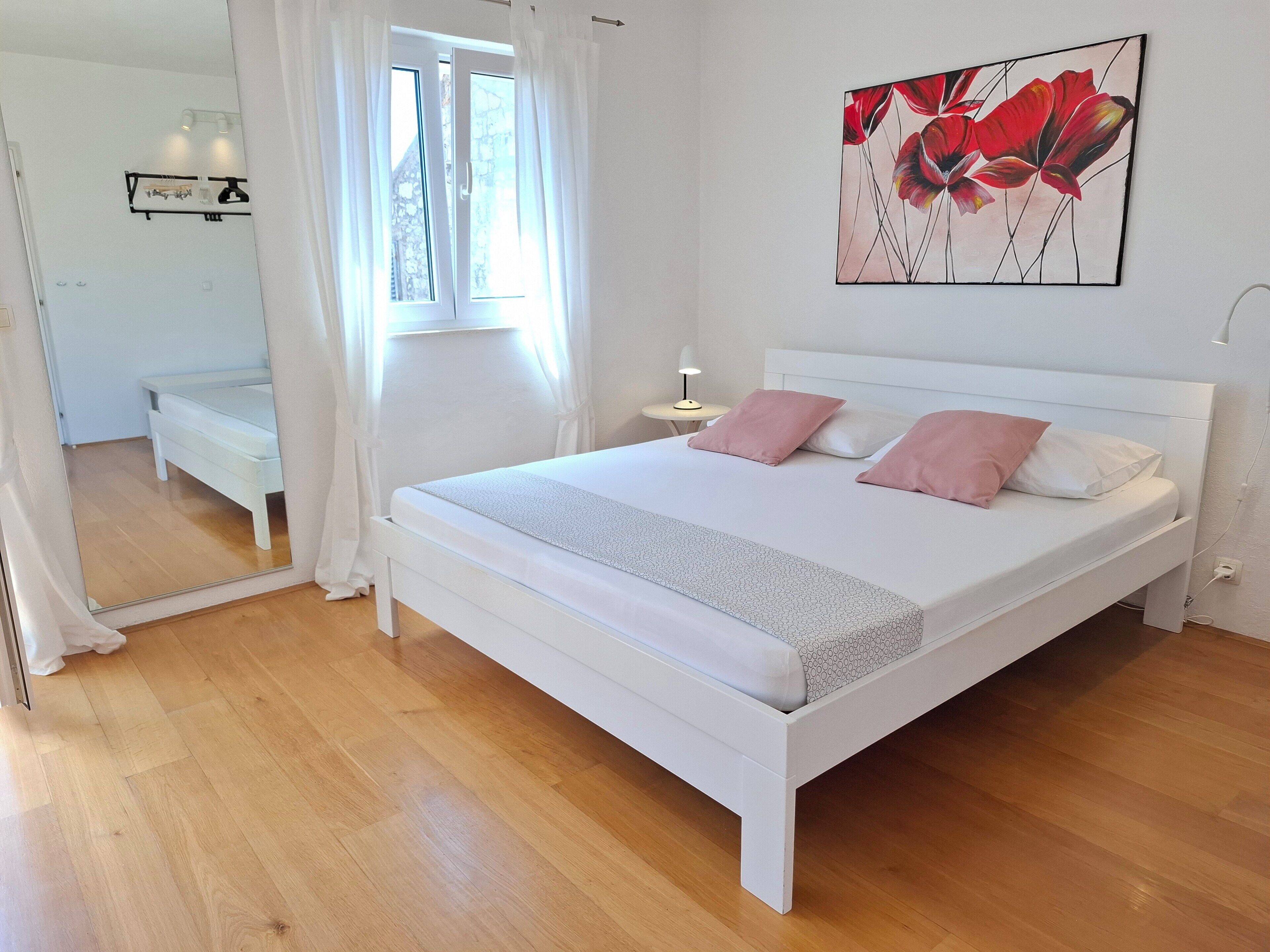 A bedroom with a white bed, pink pillows, a large mirror, and a painting of red flowers on the wall.