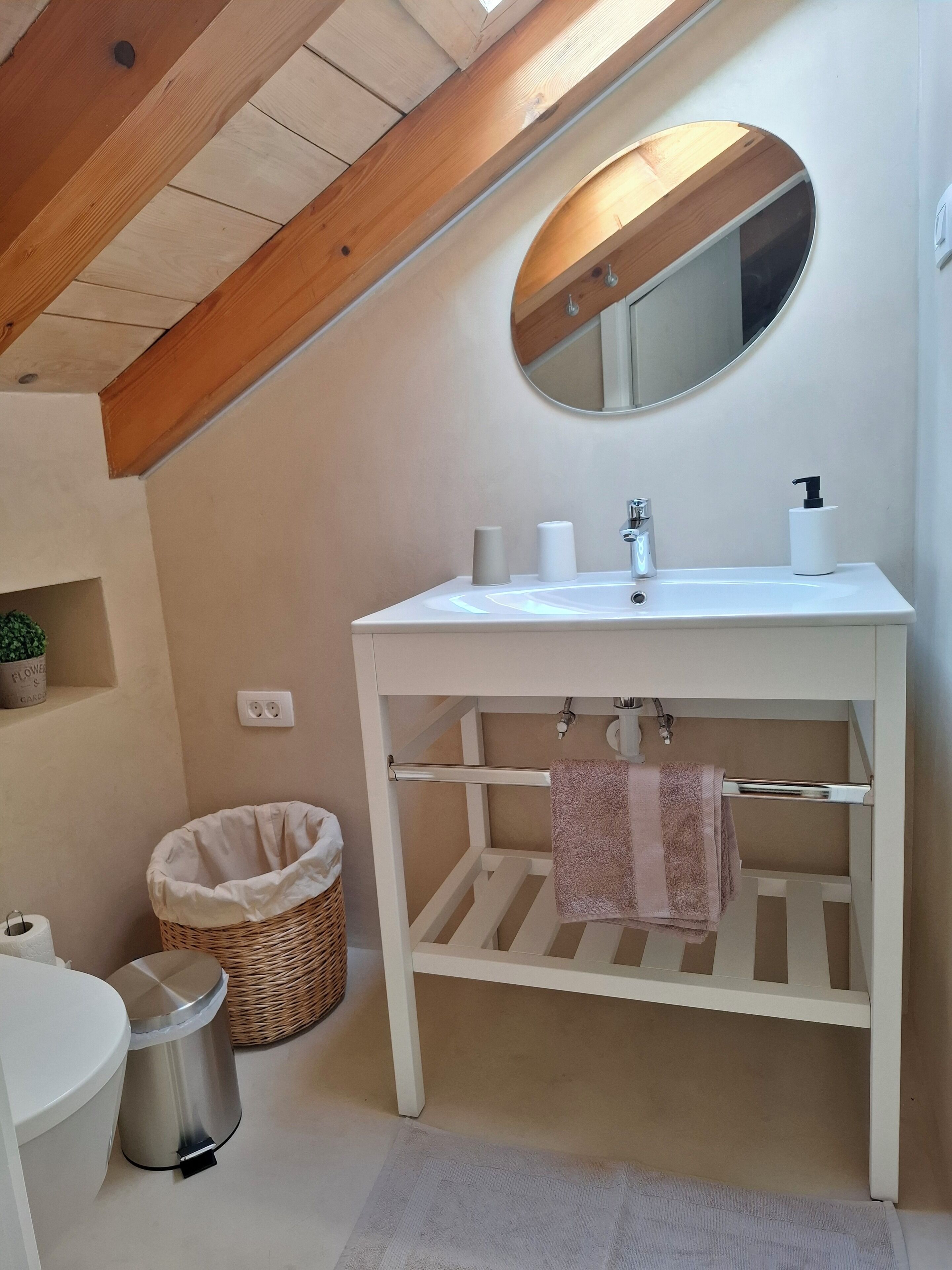 Et badeværelse med et hvidt toiletbord, et rundt spejl, en håndklædestang og en lille kurv af fletværk.