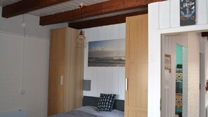 Ein Schlafzimmer mit einem Bett, Holzbalken, einer Pendelleuchte, einem an der Wand montierten Fernseher und einer Tür zu einem Flur mit einem Regal.