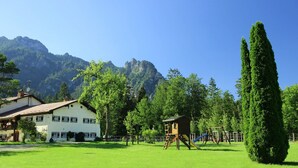 Ein Spielplatz mit Rutsche und Schaukeln, umgeben von grünem Gras und Bäumen, mit einem Berg im Hintergrund.