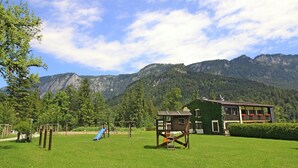 Ein Spielplatz mit Rutsche und Schaukeln, einem Holzspielhaus und einem Gebäude mit grüner Fassade und Fenstern.
