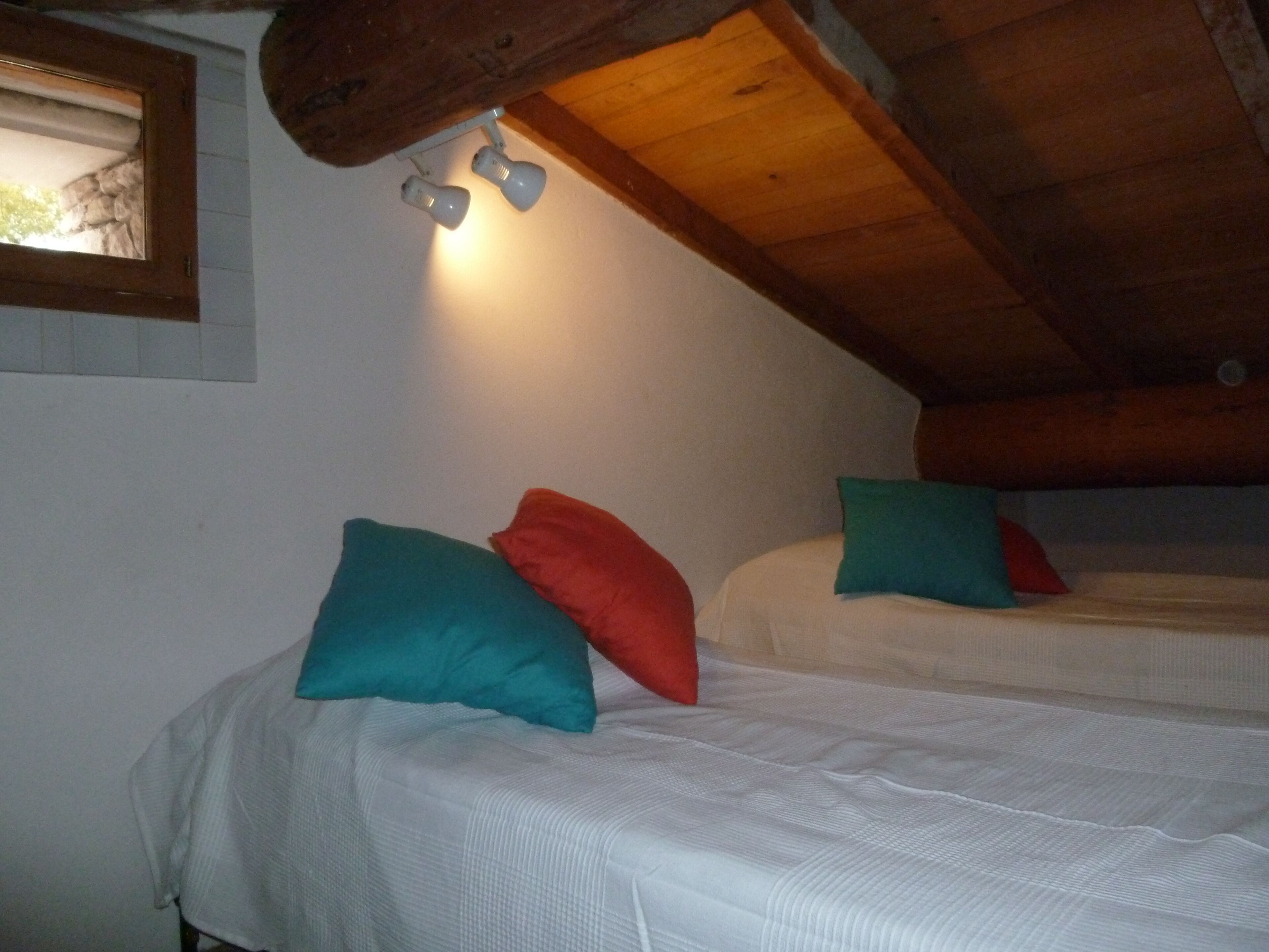 A room with two beds, each with a white mattress and teal and red pillows. There is a wooden ceiling and a window with a view of the outside.
