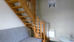 A staircase with wooden railings leading to a landing. A gray sofa is positioned below the landing, and a small round table with a white surface is visible to the right. - Charming stone cottage -air conditioner. Shared pool with 2 houses 10x5 m (Gordes)