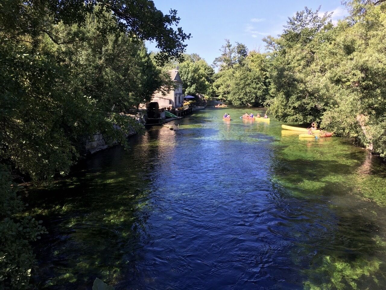 A river with kayaks, surrounded by trees and a building in the background.