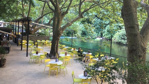An outdoor dining area with yellow chairs and tables, surrounded by trees and a river. - Charming stone cottage -air conditioner. Shared pool with 2 houses 10x5 m (Gordes)