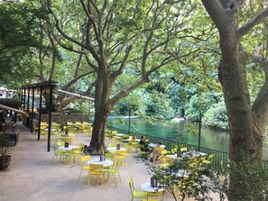 Zona pranzo all'aperto con sedie e tavoli gialli, immersa tra alberi e un fiume.