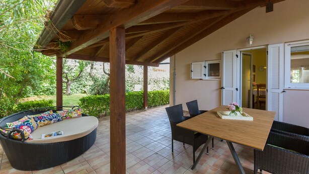 Une terrasse couverte avec une table et des chaises en bois, un canapé en osier agrémenté de coussins et une vue sur le jardin.