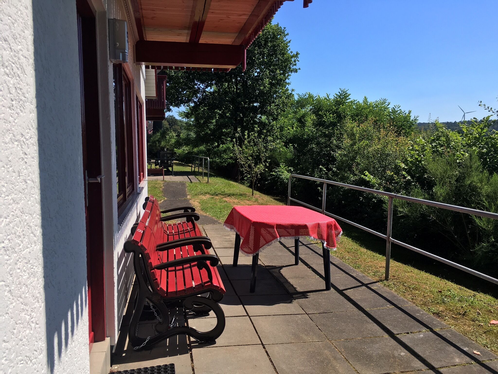 A patio with a red bench and a table, a building entrance, and a pathway leading to a green area with trees.