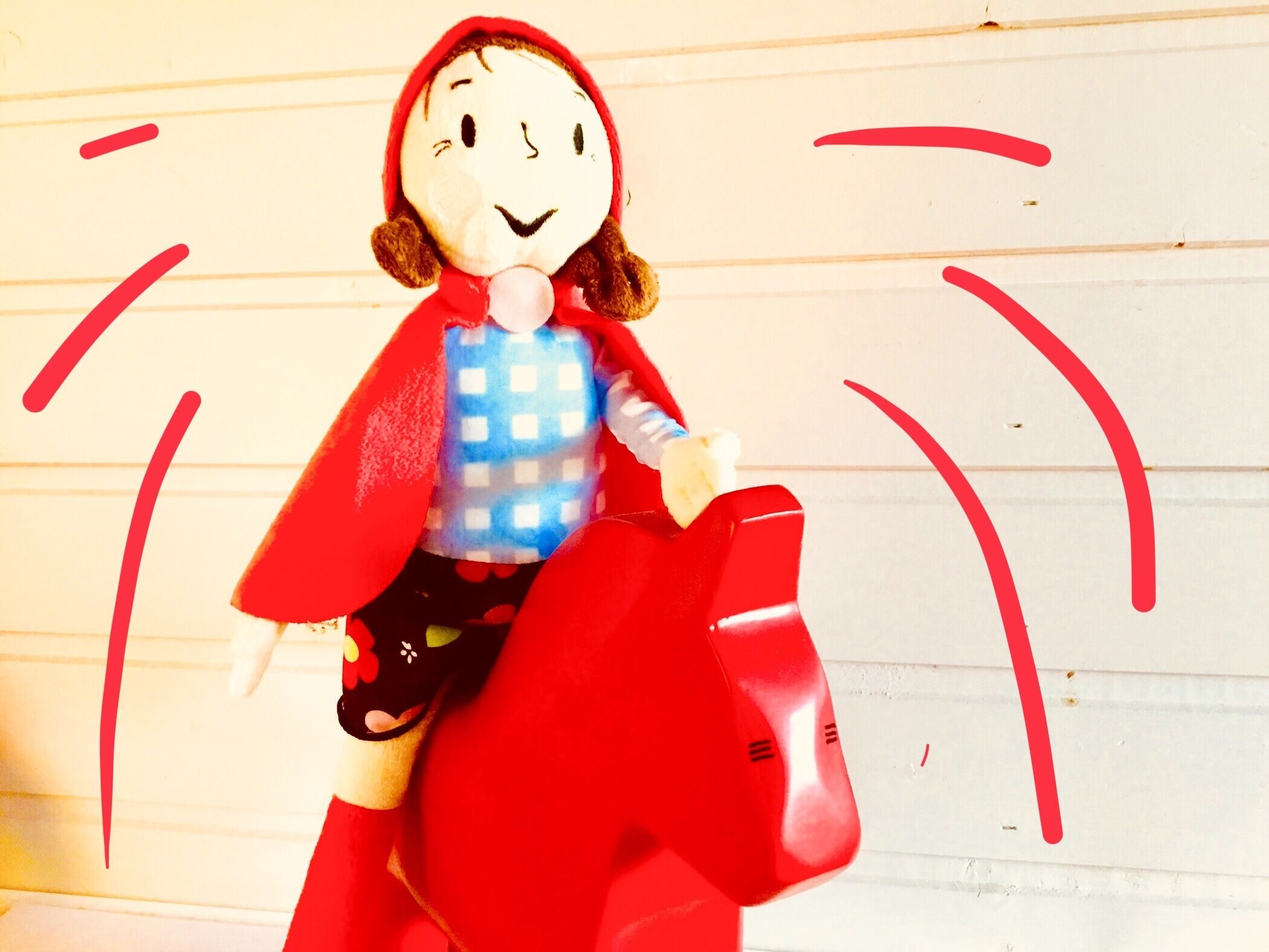 A doll with a red cape and a blue shirt riding a red horse.